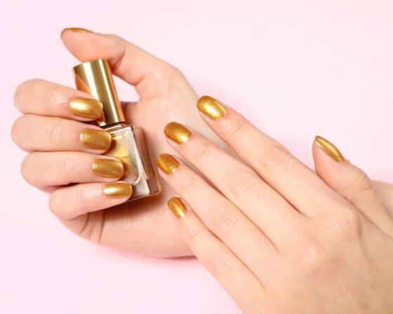 A hand holding a gold-capped bottle against a pale pink background, showing short metallic gold nails