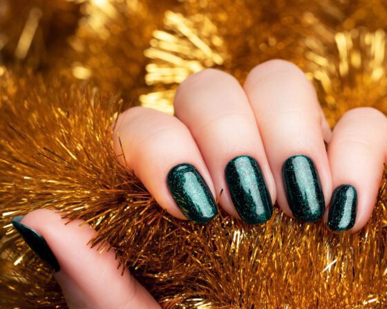 Close-up of one hand nestled in gold tinsel, showing short nails in a deep teal green with fine sparkle