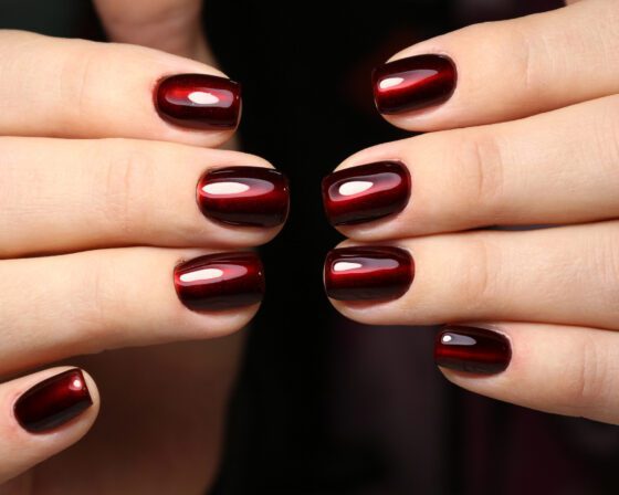 Close crop of crossed hands against dark fabric, showing short squared nails in black cherry with a luminous red centre