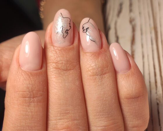 Macro of a hand against pale wood, showing glossy pale pink almond nails with silver shimmer accents and fine black line drawings