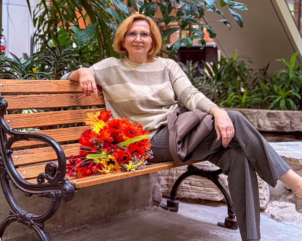 Olga Palamarenco sitting on a wooden bench in a plant-filled atrium, a bouquet of orange flowers beside her.